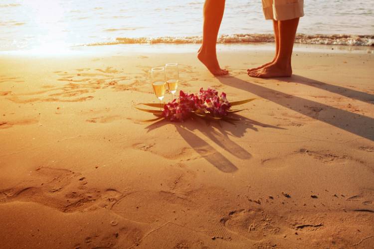 view of two people's feet, one on tip toes, and flowers in the foreground as the sunsets in the background on the beach