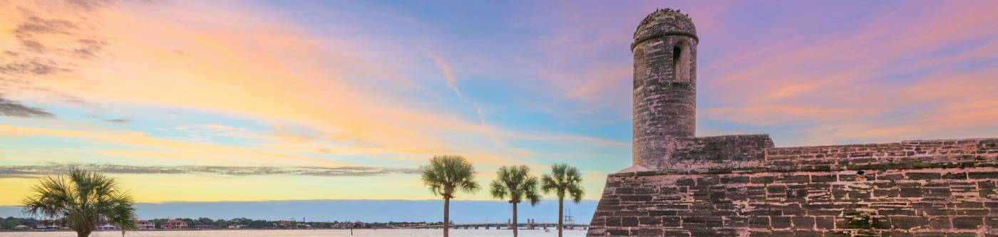 purple, blue, yellow sky over castillo de san marcos in st augustine florida with four palm trees along the water